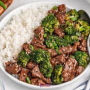 A plate with Slow Cooker Beef Broccoli stir-fry graces one side, white rice on the other. A spoon rests in the dish while sesame seeds sprinkle the top, enhancing its appeal.
