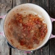 A pot on a stovetop contains sautéing onions and spices, emitting steam reminiscent of a classic calabrese pasta dish. The pot, adorned with pink handles, rests gracefully on a wooden surface.