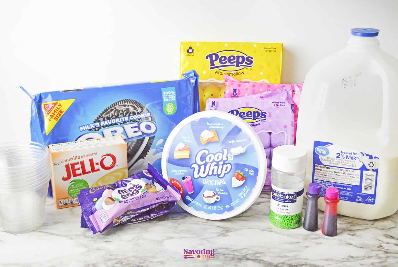 Assorted ingredients for Easter dirt cups, including Oreos, Peeps, Cool Whip, Jell-O, milk, sprinkles, food colorings, disposable cups, and chocolate chips are displayed on a marble surface.