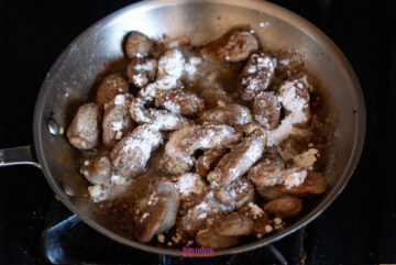 A frying pan with pieces of meat being cooked, dusted with flour, on a stovetop.
