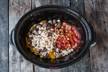 A slow cooker sausage casserole brimming with ground meat, succulent white beans, juicy tomatoes, and vibrant sliced bell peppers rests invitingly on a rustic wooden surface.