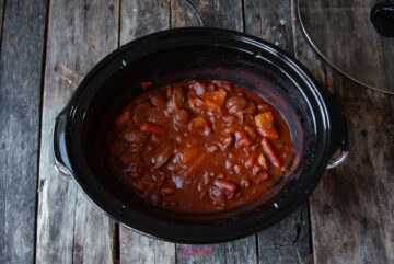 A black slow cooker filled with a hearty sausage and bean casserole in a rich red sauce, set on a rustic wooden surface.