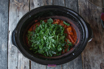 A slow cooker filled with a hearty sausage casserole, complemented by fresh chopped greens, sits invitingly on a rustic wooden surface.