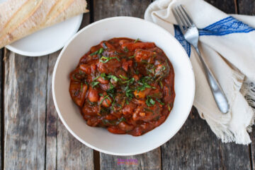 A bowl of slow-cooker vegetable stew garnished with herbs sits on a rustic wooden table, beside cutlery on a cloth napkin and a plate with sliced bread.