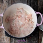 Raw chicken pieces being cooked in a pink pot on a wooden surface. A wooden spoon rests in a nearby white bowl.