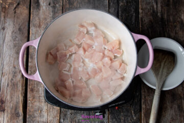 Raw chicken pieces being cooked in a pink pot on a wooden surface. A wooden spoon rests in a nearby white bowl.