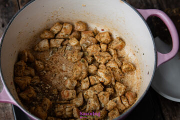Cubed chicken pieces seasoned with spices are cooked in a large, pink-handled pot, set on a wooden surface.