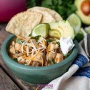 A bowl of McCormick white chicken chili is topped with shredded cheese, sour cream, sliced avocado, lime wedges, and tortilla chips. A spoon and a ripe avocado rest nearby on the rustic wooden surface.