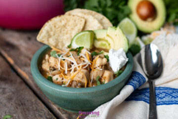 A bowl of McCormick white chicken chili is topped with shredded cheese, sour cream, sliced avocado, lime wedges, and tortilla chips. A spoon and a ripe avocado rest nearby on the rustic wooden surface.