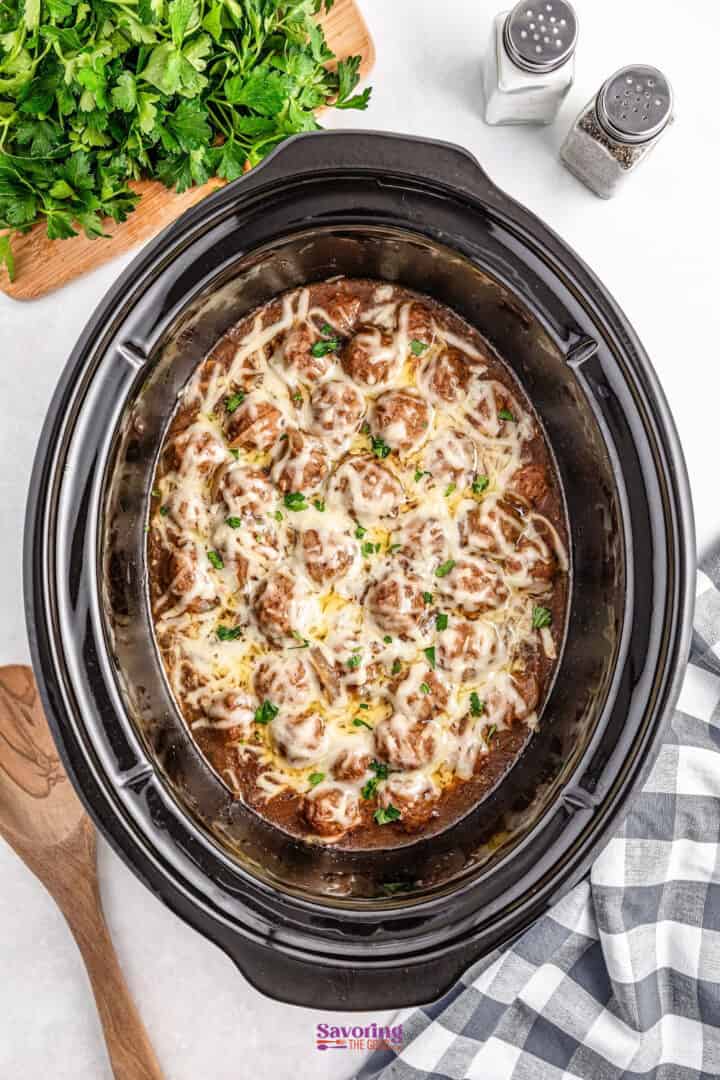 A slow cooker filled with meatballs in sauce, topped with melted cheese and garnished with herbs. Salt and pepper shakers, parsley, and a wooden spoon are nearby.