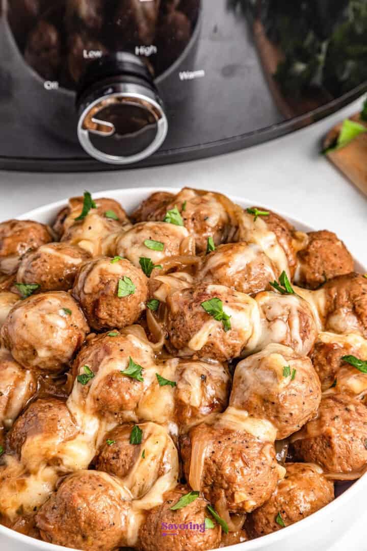 A white bowl of meatballs with melted cheese and parsley garnish is placed in front of a slow cooker.