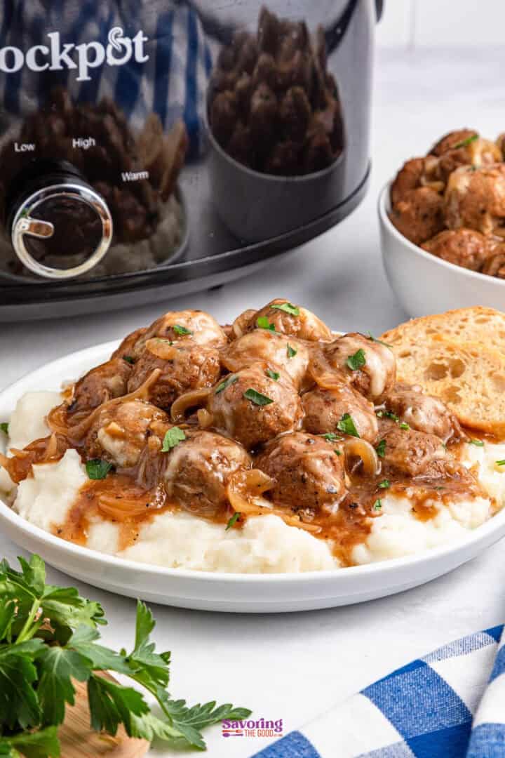 Plate of meatballs with gravy over mashed potatoes, garnished with herbs, beside a slow cooker and slice of bread on a table with a blue plaid cloth.