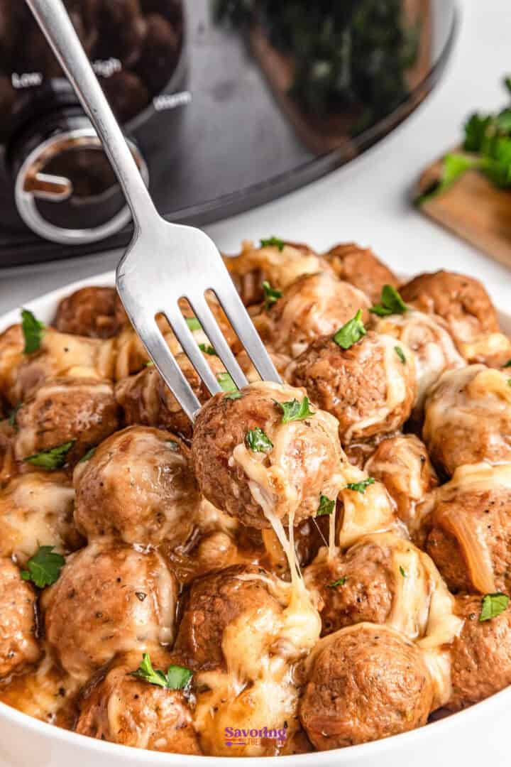 A fork lifting a cheesy meatball from a bowl filled with meatballs covered in melted cheese, with a slow cooker in the background.