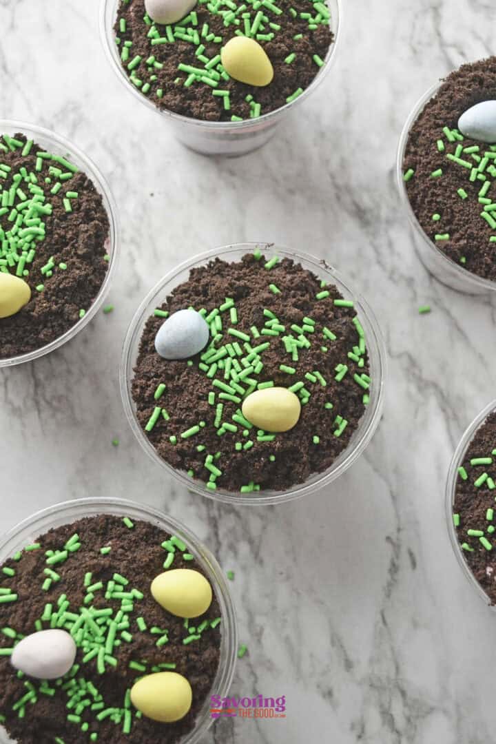 Easter dirt cups featuring plastic containers of chocolate crumbs and green sprinkles, crowned with pastel-colored candy eggs, are artfully arranged on a marble surface.