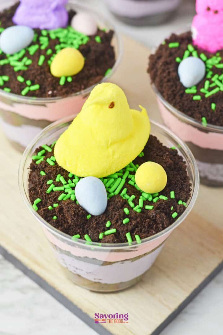 Easter dirt cups with layers of chocolate and strawberry pudding are topped with crushed cookies, green sprinkles, colorful candy eggs, and a cheerful yellow marshmallow Peep.