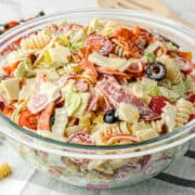 A glass bowl filled with Antipasto Pasta Salad, featuring rotini, sliced pepperoni, cubes of cheese, black olives, lettuce, cherry tomatoes, and creamy dressing.