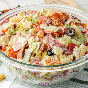 A glass bowl filled with Antipasto Pasta Salad, featuring rotini, sliced pepperoni, cubes of cheese, black olives, lettuce, cherry tomatoes, and creamy dressing.