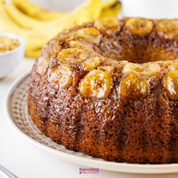 A banana cake using cake mix is topped with caramelized banana slices and sits on a plate, with fresh bananas and a bowl of brown sugar in the background.