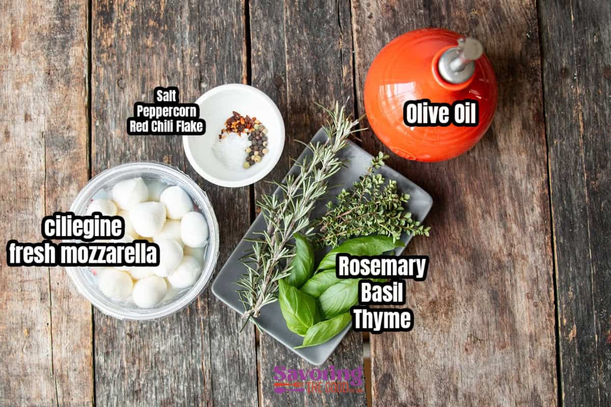 Ingredients on a wooden table: a bowl of fresh mozzarella balls, a small dish of salt, peppercorn, and chili flakes, olive oil in a bottle, sprigs of rosemary, basil, and thyme.