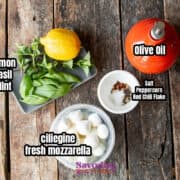 Top-down view of ingredients on a wooden surface, featuring marinated mozzarella balls on a plate, surrounded by lemon, basil, mint, olive oil in a jug, and spices in a bowl.