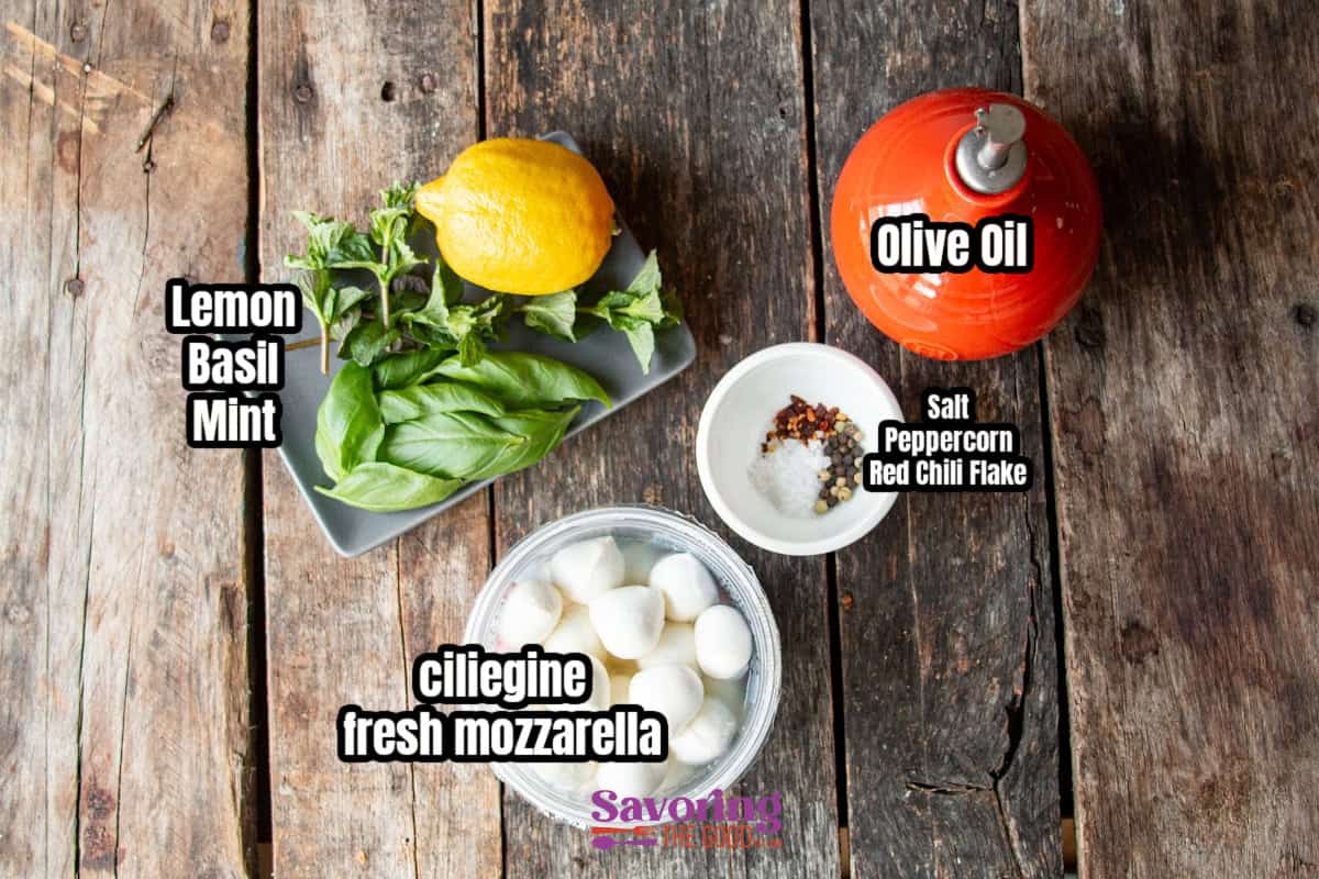 Top-down view of ingredients on a wooden surface, featuring marinated mozzarella balls on a plate, surrounded by lemon, basil, mint, olive oil in a jug, and spices in a bowl.