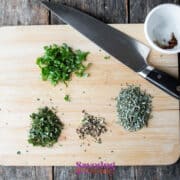Chopped herbs and spices on a wooden cutting board with a knife. Nearby, a small bowl contains salt and pepper flakes.