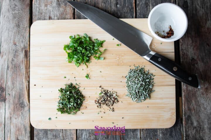Chopped herbs and spices on a wooden cutting board with a knife. Nearby, a small bowl contains salt and pepper flakes.