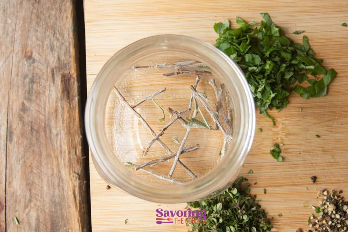 Empty glass jar on a wooden surface, surrounded by chopped herbs and spices, with some herb stems inside.