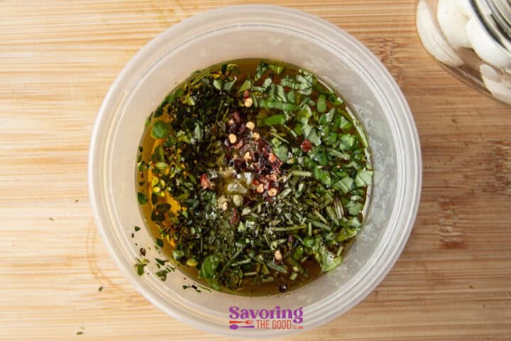 A plastic container filled with olive oil, chopped herbs, and red pepper flakes. A glass jar is partially visible on the wooden surface.