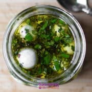 Top view of a glass jar containing marinated mozzarella balls in olive oil, herbs, and spices. A spoon is partially visible beside the jar on a wooden surface.