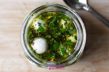 Top view of a glass jar containing marinated mozzarella balls in olive oil, herbs, and spices. A spoon is partially visible beside the jar on a wooden surface.