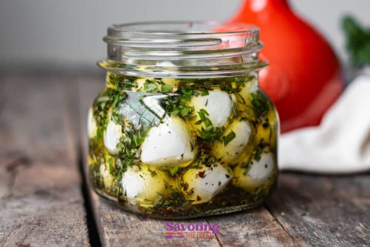 A jar of marinated mozzarella balls with herbs and oil sits on a wooden surface, accompanied by a red object in the background.