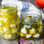 Two jars of marinated cheese sit on a wooden surface. One jar contains yellow marinated cubes, while the other holds marinated mozzarella balls in oil with herbs. A spoon rests in the open jar, ready to serve these delightful bites.