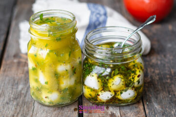 Two jars of marinated cheese sit on a wooden surface. One jar contains yellow marinated cubes, while the other holds marinated mozzarella balls in oil with herbs. A spoon rests in the open jar, ready to serve these delightful bites.