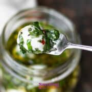 A fork holding a marinated mozzarella ball garnished with chopped herbs, above a jar filled with more mozzarella balls and oil.