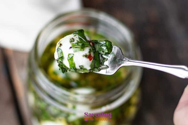 A fork holding a marinated mozzarella ball garnished with chopped herbs, above a jar filled with more mozzarella balls and oil.
