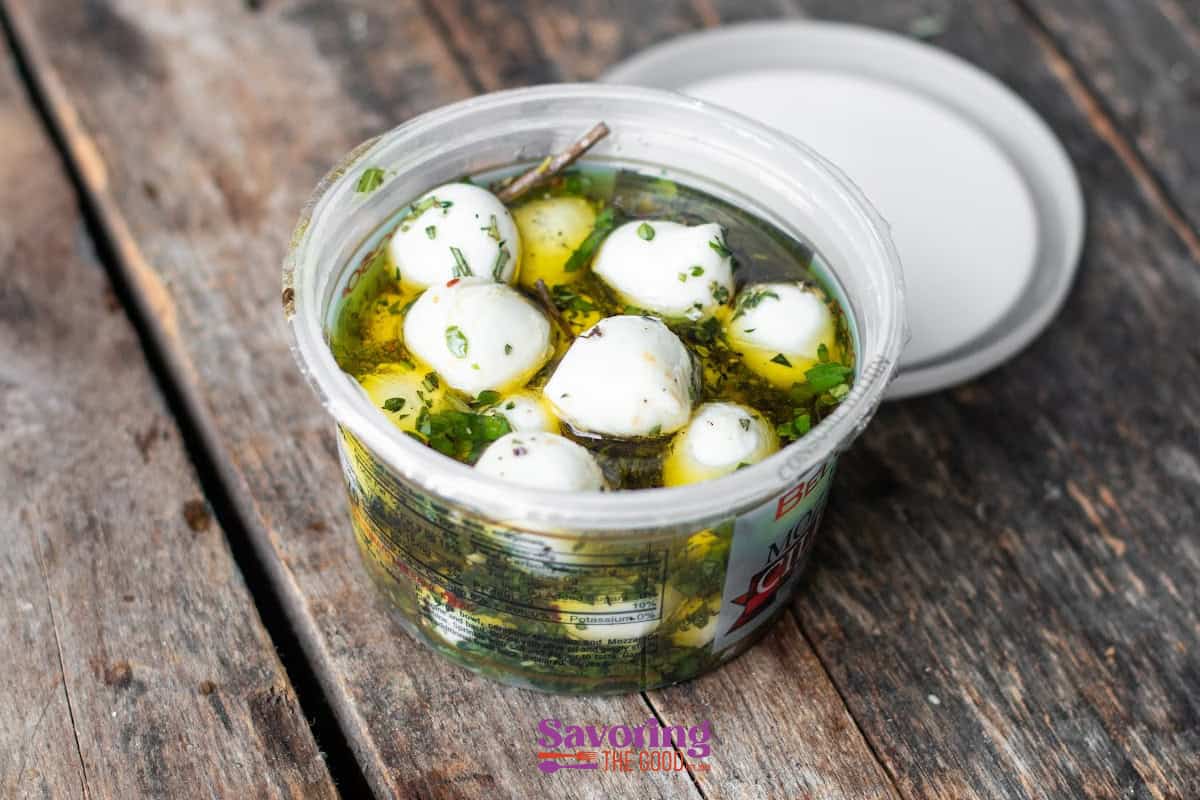 A plastic container of marinated mozzarella balls with herbs and oil sits on a rustic wooden surface, lid placed beside it.
