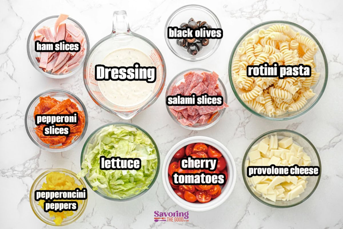 Various ingredients for an Italian pasta salad are arranged in bowls, including rotini pasta, ham, salami, pepperoni, lettuce, provolone cheese, cherry tomatoes, olives, peppers, and dressing.