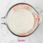 A glass measuring cup filled with a creamy white sauce containing small black specks, perfect as an Antipasto Pasta Salad dressing, sits on a white marble surface.