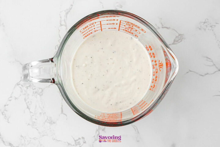 A glass measuring cup filled with a creamy white sauce containing small black specks, perfect as an Antipasto Pasta Salad dressing, sits on a white marble surface.