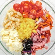 A glass bowl filled with Antipasto Pasta Salad-rotini, cherry tomatoes, pepperoni, salami, mozzarella cubes, black olives, banana pepper rings, and sliced cheese arranged in colorful sections.