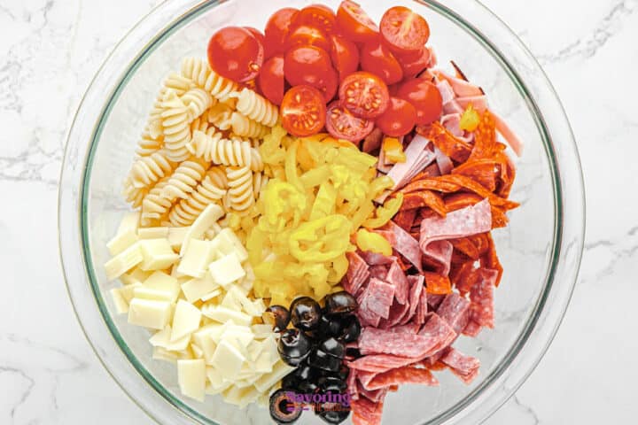 A glass bowl filled with Antipasto Pasta Salad-rotini, cherry tomatoes, pepperoni, salami, mozzarella cubes, black olives, banana pepper rings, and sliced cheese arranged in colorful sections.