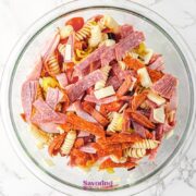 A glass bowl filled with Antipasto Pasta Salad-rotini pasta, sliced deli meats, cheese cubes, and vegetables-sits on a marble surface.