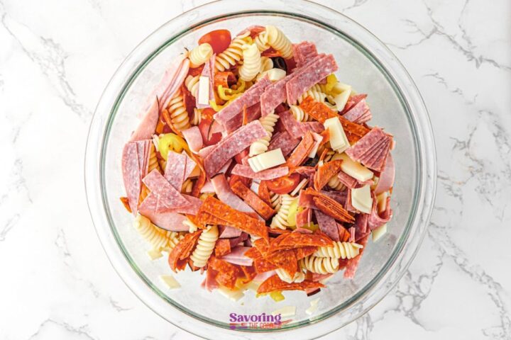A glass bowl filled with Antipasto Pasta Salad-rotini pasta, sliced deli meats, cheese cubes, and vegetables-sits on a marble surface.
