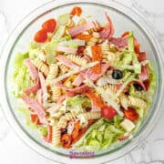 A glass bowl filled with Antipasto Pasta Salad featuring rotini pasta, lettuce, sliced deli meats, cherry tomatoes, black olives, and cheese on a white marble surface.