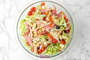 A glass bowl filled with Antipasto Pasta Salad featuring rotini pasta, lettuce, sliced deli meats, cherry tomatoes, black olives, and cheese on a white marble surface.