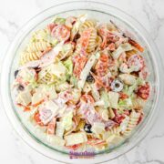 A glass bowl filled with creamy Antipasto Pasta Salad, featuring rotini, cherry tomatoes, bell peppers, olives, cheese cubes, and sliced meats on a white marble surface.