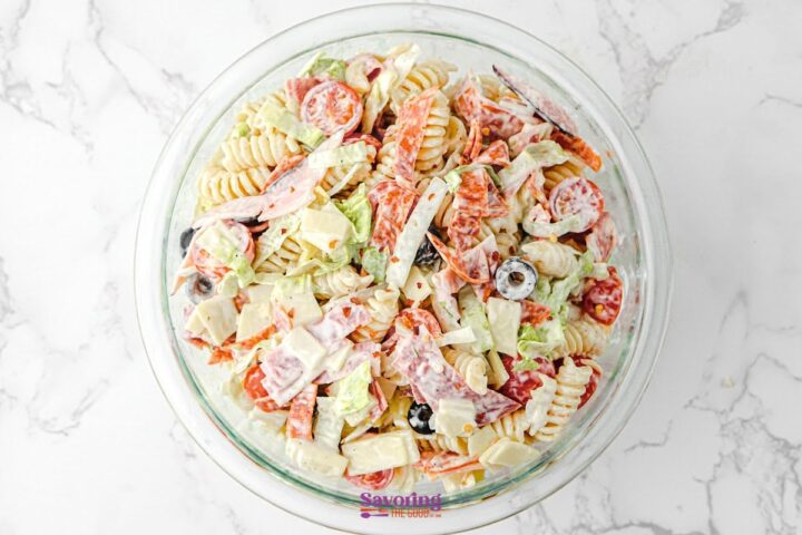 A glass bowl filled with creamy Antipasto Pasta Salad, featuring rotini, cherry tomatoes, bell peppers, olives, cheese cubes, and sliced meats on a white marble surface.