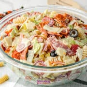 A glass bowl filled with Antipasto Pasta Salad-rotini, lettuce, cherry tomatoes, olives, cheese, and sliced meats-rests on a striped cloth with tomatoes and utensils in the background.