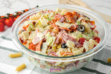 A glass bowl filled with Antipasto Pasta Salad—rotini, lettuce, cherry tomatoes, olives, cheese, and sliced meats—rests on a striped cloth with tomatoes and utensils in the background.